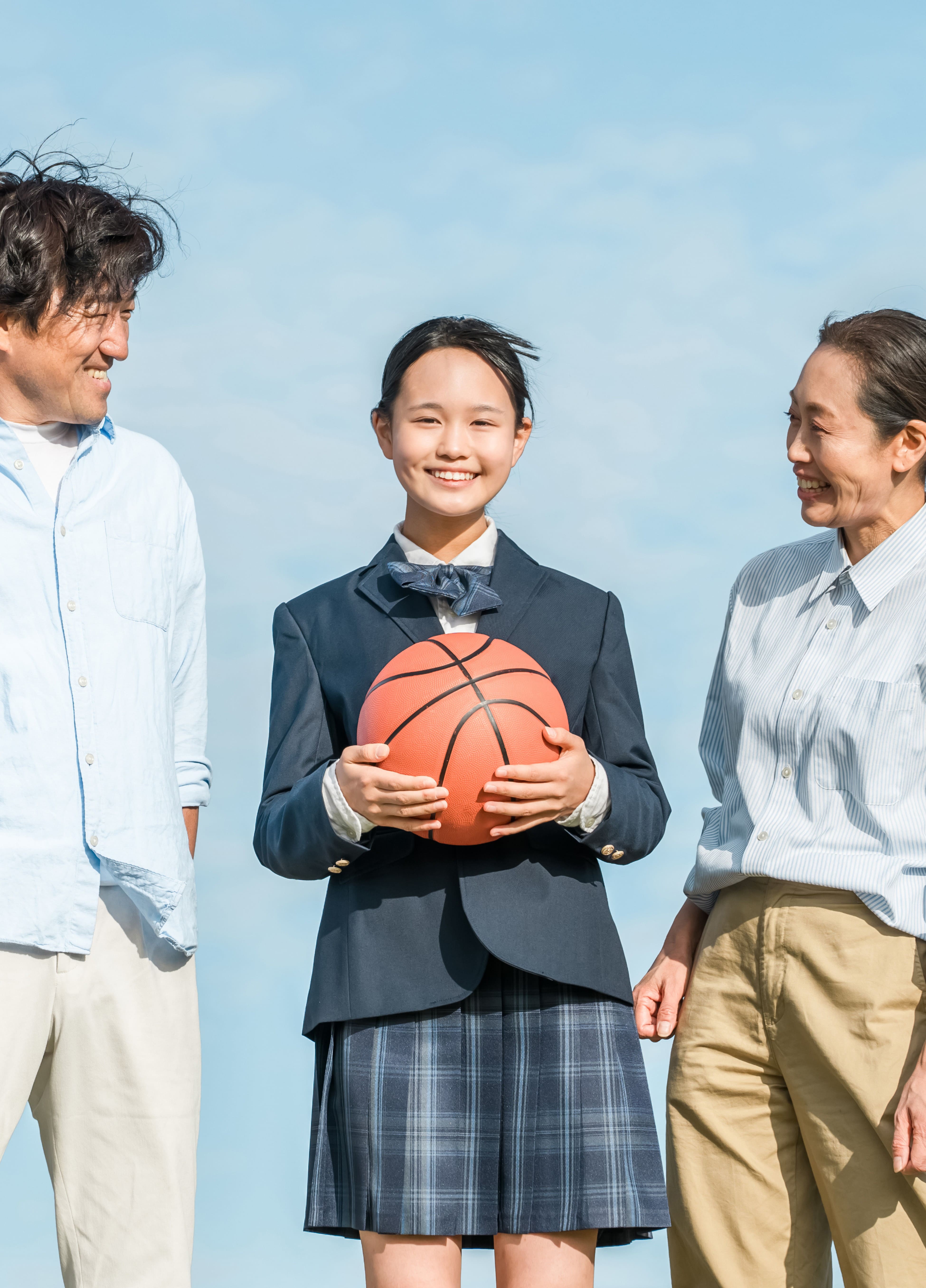 制服姿の女子生徒がバスケットボールを持ち、保護者らしき大人と笑顔で並んでいる。