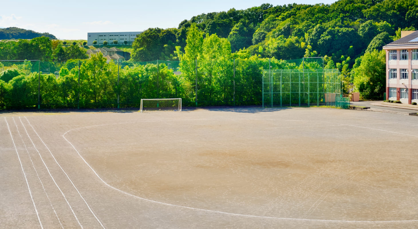 緑に囲まれた中学校の広いグラウンド。奥には校舎と山が見える。